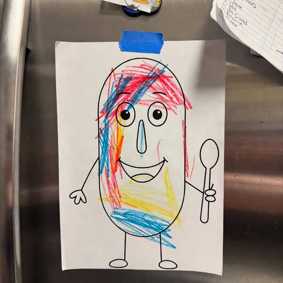 Child's crayon drawing of smiling bean holding spoon on fridge