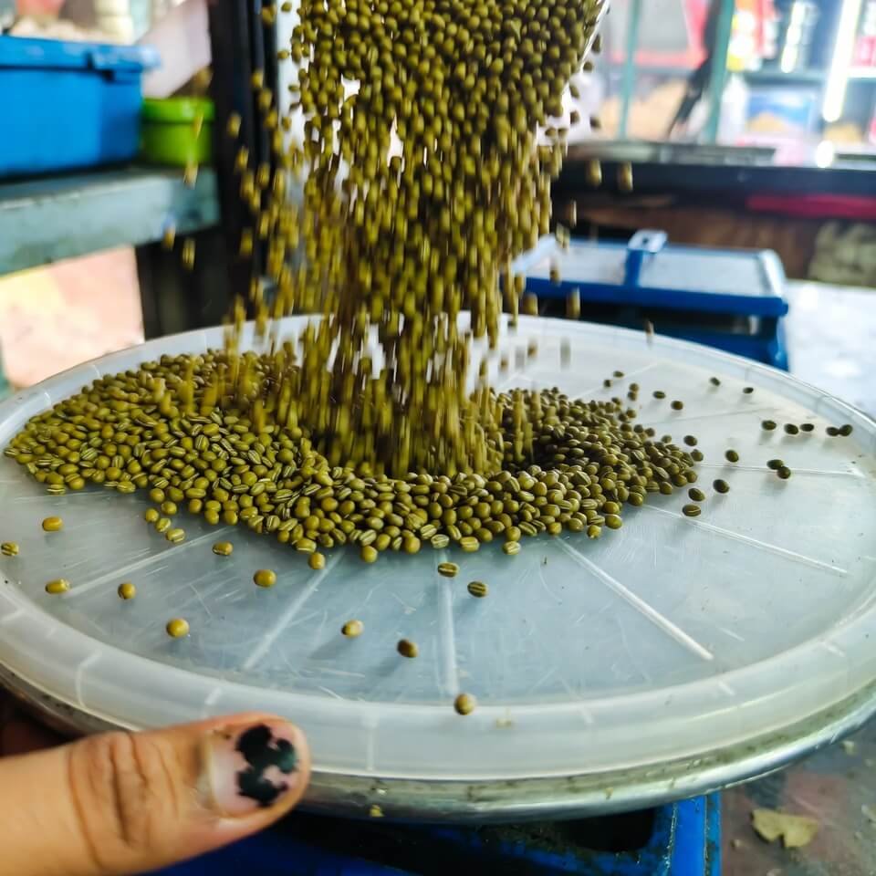 Hand pouring green moong dal into scratched container on market scale.