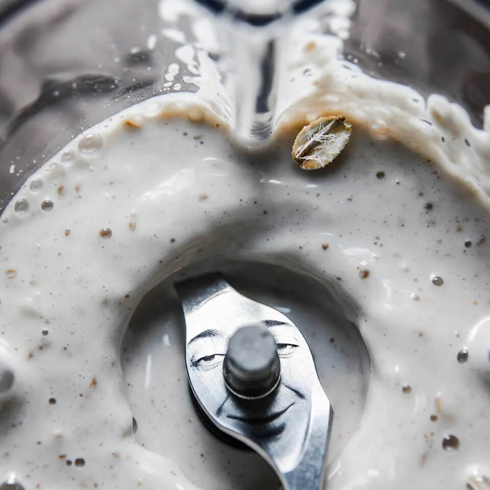 Blender jar with swirling oat pulp and tired face reflection.